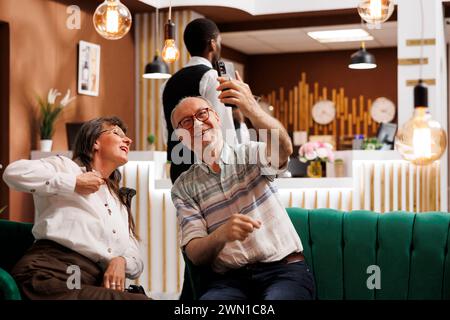 Vista dalla lussuosa hall dell'hotel dei turisti anziani in pensione che realizzano videocassette Internet. Felice coppia anziana in luna di miele, in contatto con la famiglia e gli amici tramite telefono cellulare. Foto Stock