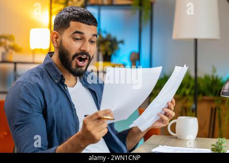 Uomo con la barba indiana libero professionista che lavora con documenti dall'ufficio domestico, analizzando le informazioni, leggendo documenti aziendali preparano un rapporto finanziario. Giovane contabile arabo che analizza documenti grafici. Foto Stock