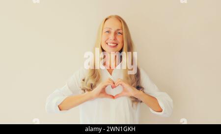 Ritratto di una bella donna di mezza età sorridente e felice che mostra un gesto a forma di cuore con le mani sul petto su sfondo bianco dello studio Foto Stock