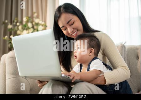 Una felice donna d'affari asiatica lavora da casa, lavora sul suo portatile e si prende cura del suo bambino cattivo. Foto Stock