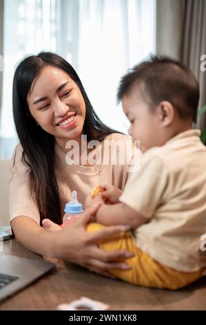 Una mamma asiatica felice sta giocando e nutrendo il suo bambino con spuntini mentre lavora nel suo ufficio domestico. lavora da casa e dai concetti di vita della mamma Foto Stock