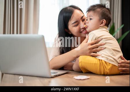 Una mamma asiatica felice sta giocando e nutrendo il suo bambino con spuntini mentre lavora nel suo ufficio domestico. lavora da casa e dai concetti di vita della mamma Foto Stock