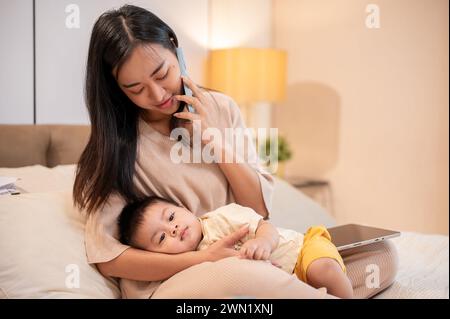 Una mamma asiatica premurosa sta mettendo il suo bambino a dormire tra le braccia mentre parla al telefono con qualcuno. pisolino e buonanotte, maternità e.. Foto Stock