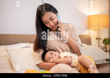 Una mamma asiatica premurosa sta mettendo il suo bambino a dormire tra le braccia mentre parla al telefono con qualcuno. pisolino e buonanotte, maternità e.. Foto Stock