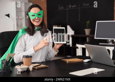Donna d'affari vestita da supereroina che indica un tablet in ufficio Foto Stock