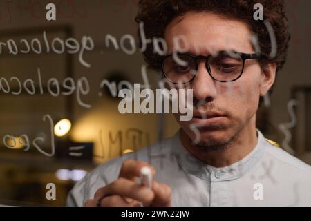 Primo piano di un giovane uomo che indossa occhiali da vista che sviluppa codice informatico che lo scrive su una parete di vetro Foto Stock