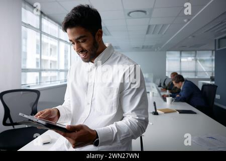 Giovane uomo d'affari sicuro di sé in abbigliamento da lavoro sorridente e che utilizza un tablet digitale in ufficio Foto Stock