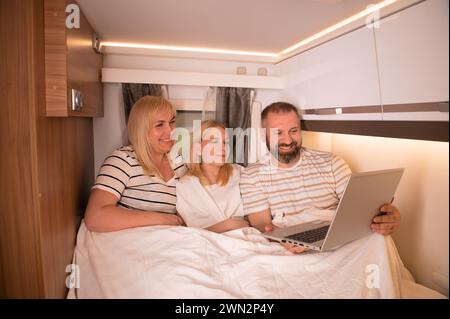 Una famiglia di tre persone sta guardando un film su un computer portatile mentre si siede sul letto del loro camper Foto Stock