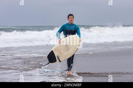 Un surfista stanco e bagnato con una muta blu esce dall'oceano tenendo la tavola da surf, con le onde che si schiantano dietro di lui. Foto Stock