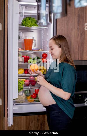 Giovane donna incinta con un'arancia vicino al frigorifero di casa. Mangiare sano. Foto Stock