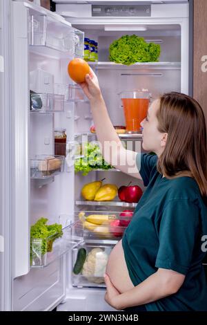 Giovane donna incinta con un'arancia vicino al frigorifero di casa. Mangiare sano. Foto Stock