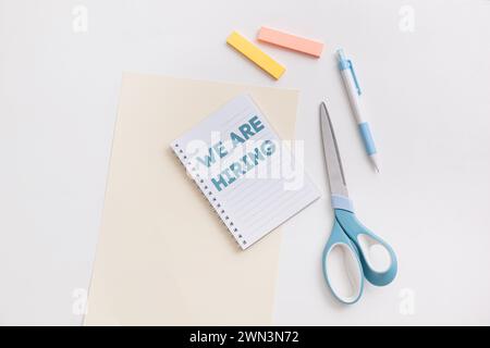 Una scrivania bianca con scritto We Are Hiring, con un quaderno blu di forbici, penna, evidenziatori, che simboleggia le opportunità di lavoro e. Foto Stock