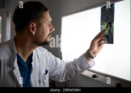 Vista laterale di un medico serio che studia i risultati della risonanza magnetica. Specialista maschile che lavora in ospedale, effettuando la descrizione delle radiografie del paziente, puntando da penna. Concetto di medicina e ospedale. Foto Stock