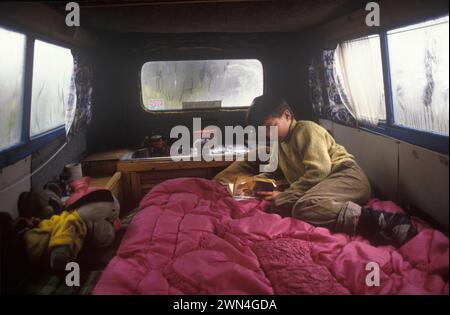 OFF Grid Living 1980s UK. Bambino, giovane ragazzo nel furgone del padre che legge un libro. (Watership Down) Kevin Oubridge viveva con la sua famiglia nella valle di tipi, una comunità hippy gallese che ospitava circa 120 spiriti liberi eco-compatibili. Llandeilo, Galles 1985 HOMER SYKES Foto Stock