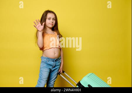Adorabile bambina viaggiatrice che porta la valigia blu, saluta con la mano, guarda la macchina fotografica, isolata sullo sfondo giallo dello studio. Copia pubblicità Foto Stock