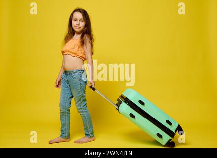 Foto a grandezza naturale di un'adorabile bambina viaggiatrice con la parte superiore arancione e il denim blu, che trasporta una elegante valigia blu, cammina a piedi nudi lungo un bordello giallo Foto Stock