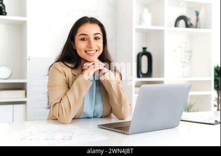 Ritratto di una bruna indiana o araba di successo con un abito elegante, dipendente aziendale, seduta in un ufficio moderno, guardando la macchina fotografica e sorridente Foto Stock