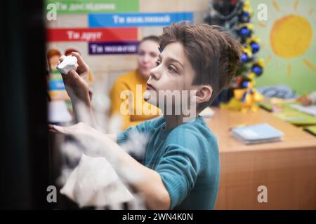 Charkiv, Ucraina. 19 dicembre 2023. Un ragazzo scrive sulla lavagna durante una lezione di classe con il permesso del suo insegnante. I bambini ucraini frequentano le lezioni in una scuola in una stazione della metropolitana di Charkiv, Ucraina, nel mezzo dell'invasione russa. Molti bambini a Charkiv non vanno a scuola perché i loro genitori temono per la loro sicurezza a causa della vicinanza della città al confine russo. Le autorità cittadine hanno organizzato il processo di istruzione per i bambini nelle classi della scuola sotterranea nella metropolitana della città. (Credit Image: © Mykhaylo Palinchak/SOPA Images via ZUMA Press Wire) SOLO PER USO EDITORIALE! Non per Comm Foto Stock