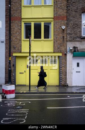 Bermondsey, Londra, Regno Unito: Una donna con un cappotto nero che cammina lungo Bermondsey Street. Passa davanti a un edificio in mattoni con i lavori in legno dipinti di giallo. Foto Stock