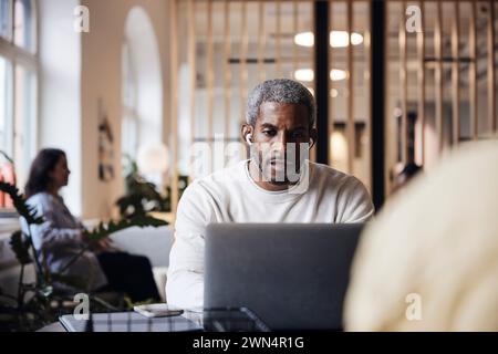 Professionisti uomini maturi che lavorano con un laptop indossando cuffie intrauricolari wireless in ufficio Foto Stock