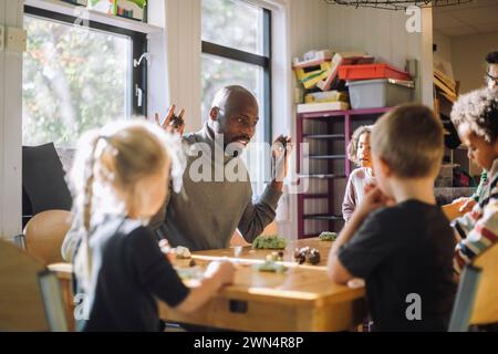 Insegnante di sesso maschile che spiega ai bambini seduti alla panchina durante la lezione di formatura dell'argilla all'asilo Foto Stock
