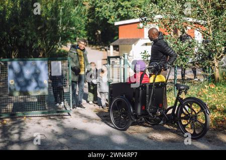 Insegnante donna in piedi al cancello mentre accoglie i bambini seduti in bici cargo all'asilo Foto Stock