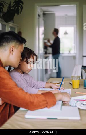Un uomo gay che assiste la figlia a fare i compiti mentre si siede al tavolo da pranzo a casa Foto Stock