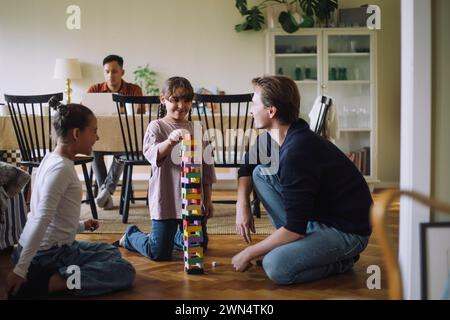 Ragazza sorridente che sta impilando i blocchi di Jenga mentre si inginocchia sul pavimento da padre e sorella a casa Foto Stock