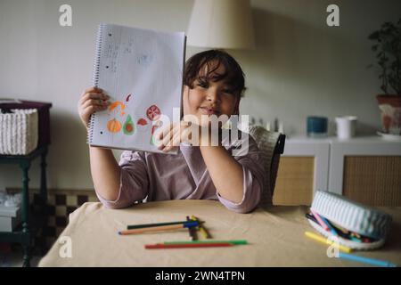 Ritratto di una ragazza sorridente che mostra il disegno su un notebook mentre è seduta a casa Foto Stock