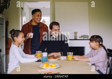 Felice coppia gay che si diverte mentre fa colazione con le figlie al tavolo da pranzo a casa Foto Stock