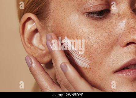Primo piano di una giovane donna che applica la crema sulla pelle lentigginata. Colpo ritagliato di donna che applica idratante sulla guancia con le dita. Foto Stock