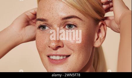 Primo piano ritratto dettagliato di una giovane donna sorridente con lentiggini. Immagine ritagliata di una bella donna bionda che regola i capelli contro un bac beige Foto Stock