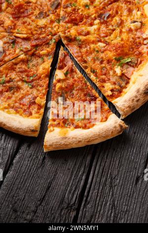 Pizza con diversi tipi di carne, tagliata a fette, una fetta è separata su uno sfondo di legno nero, vista dall'alto Foto Stock