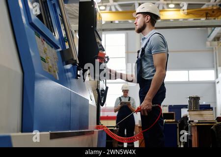 Vista laterale medio-lunga ad angolo basso dell'uomo caucasico con barba che indossa un'uniforme con elmetto utilizzando una macchina CNC al lavoro in fabbrica Foto Stock