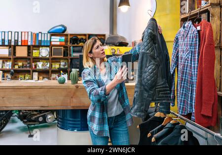 Donna bionda che tiene in mano un tablet digitale mentre controlla la giacca in pelle nera sul negozio di abbigliamento di seconda mano Foto Stock
