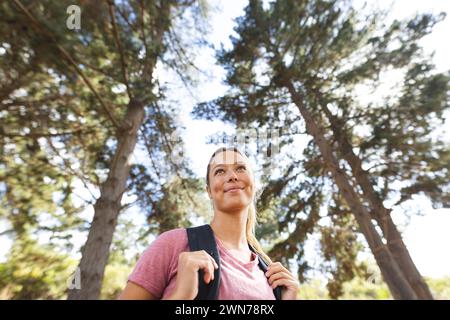 La giovane donna caucasica con uno zaino guarda con attenzione tra alberi alti durante un'escursione Foto Stock