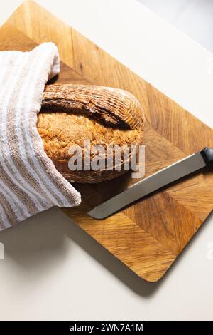 Una pagnotta di pane e un coltello poggiano su un tagliere di legno, parzialmente coperto da un asciugamano a strisce Foto Stock