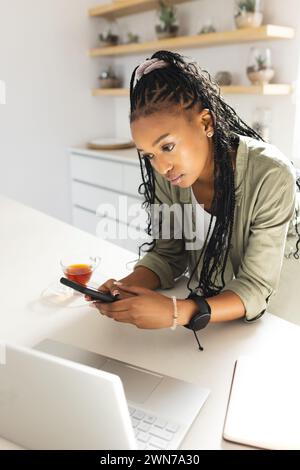 Una giovane donna afroamericana sfoglia il suo smartphone mentre tiene una tazza di tè Foto Stock