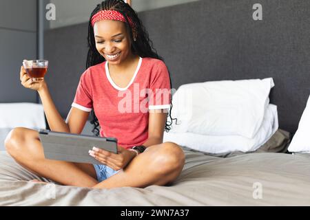 La giovane donna afroamericana si diverte con un tablet e un tè a letto Foto Stock