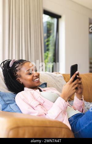 Una giovane donna afro-americana sdraiata su un divano, sorridendo al suo telefono, con spazio di copia Foto Stock