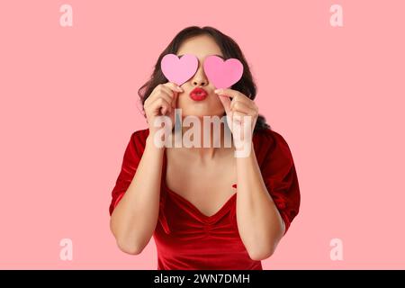 Bella giovane donna asiatica felice con cuori di carta che soffiano bacio su sfondo rosa. Festa di San Valentino Foto Stock