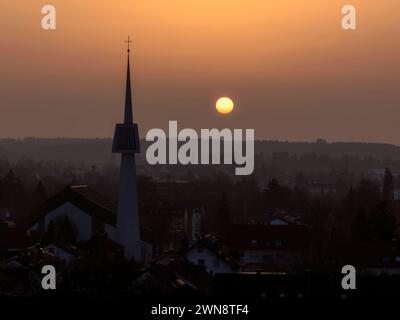 29.02.2024, Bad Wörishofen, Drohnenaufnahme. Die Sonne Geht als glutroter Ball hinter dem modernen Beton-Kirchturm von St Ulrich in der Gartenstadt unter. Der Dunst am Abendhimmel sorgt für einen Sonnenuntergang wie man ihn sonst nur von Meeresküsten kennt. Möglich macht diese seltene Aufnahme ein Teleobjektiv in einer Fotodrohne, aufgestiegen am richtigen Platz zur richtigen Zeit. 29.02.2024, Bad Wörishofen 29.02.2024, Bad Wörishofen *** 29 02 2024, Bad Wörishofen, drone ha sparato il sole tramonta come una palla rossa ardente dietro la moderna torre della chiesa di cemento di Sant'Ulrico nella città giardino la foschia i Foto Stock