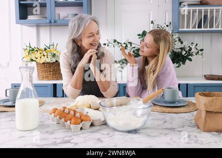 Donna anziana felice che insegna alla ragazza carina di impastare l'impasto mentre parla della vita. Unità di generazioni Foto Stock