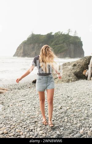 Giovane donna che cammina su una spiaggia rocciosa con lunghi capelli che soffiano nel vento Foto Stock