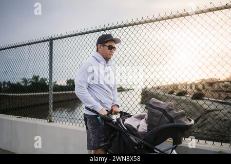 Uomo con cappello e occhiali da sole che spinge il bambino nel passeggino sul ponte Foto Stock