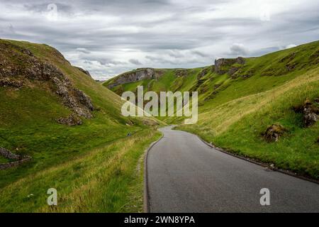 Affacciato su una strada vuota del Winnat's Pass nel Peak District National Park con due camminatori che camminano su, Derbyshire, Regno Unito Foto Stock