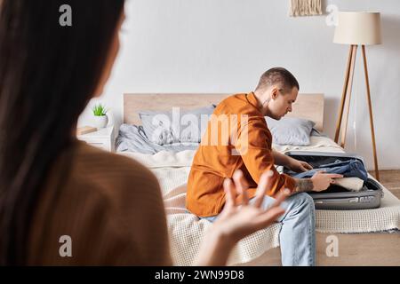 triste uomo tatuato che sta imballando la valigia vicino alla moglie litigando in camera da letto a casa, malinteso Foto Stock