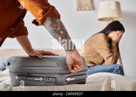 giovane uomo tatuato che sta imballando valigia vicino a moglie asiatica che piange sul letto a casa, malinteso familiare Foto Stock