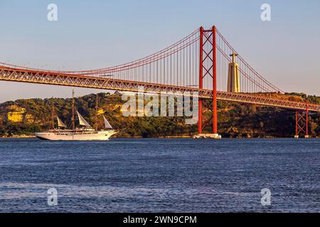 Una nave naviga sotto un grande ponte sospeso durante un sereno tramonto, riflettendo sfumature arancioni sull'acqua, Lissabon, 25 de Abril Bridge Foto Stock