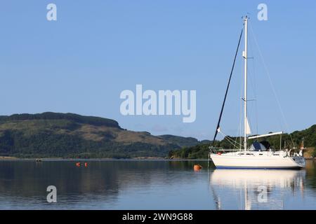 Yacht a vela bianco su un lago di mare calmo, con colline verdi che si riflettono nell'acqua liscia Foto Stock